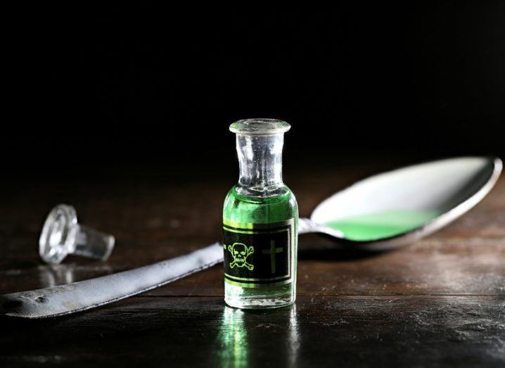 Illustration of a glass vial with green liquid inside it and a skull and crossbones image on the front. The vial is in front of a spoon that has some green liquid on it, which is on top of a dark wooden table.