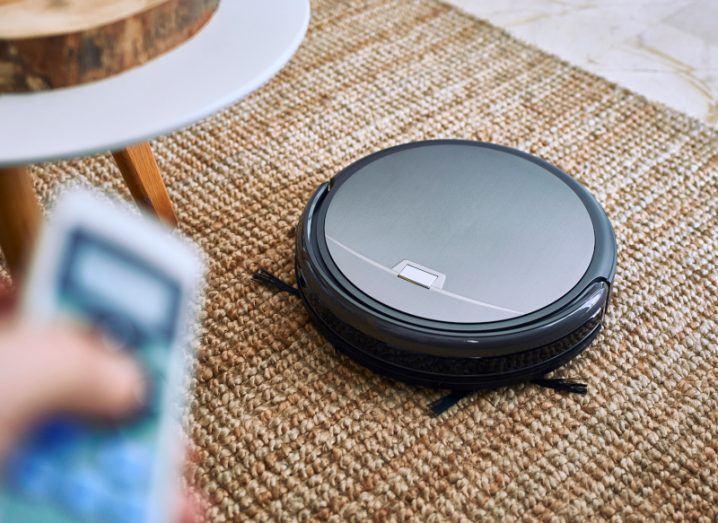 A robot vacuum cleaner on a mat in a living room. Someone is pointing a remote control at it.