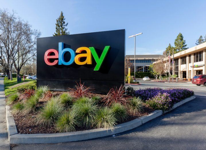 A black sign with the eBay logo on it and a building behind it. Trees and a clear blue sky are visible in the background.