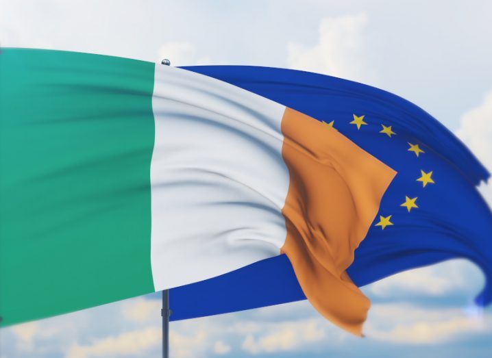 A flag of Ireland blowing in the wind with an EU flag behind it and clouds in the background.