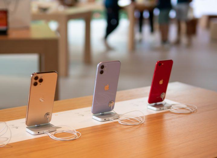 Three Apple iPhones on a wooden table, with the Apple logo visible on the back of each smartphone. There are other tables and people walking in the background.