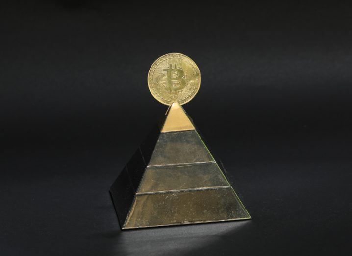 Illustration of a pyramid with a gold bitcoin on the top of it, in a black background.
