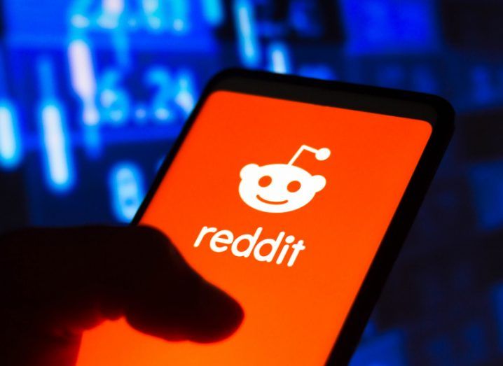 The Reddit logo on a smartphone screen. The phone is in a person's hand and there are blue numbers floating in the background of the image.