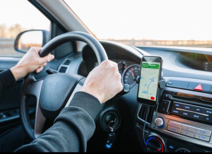 Person's hands on a steering wheel driving a car with a smartphone with a GPS device beside the steering wheel.