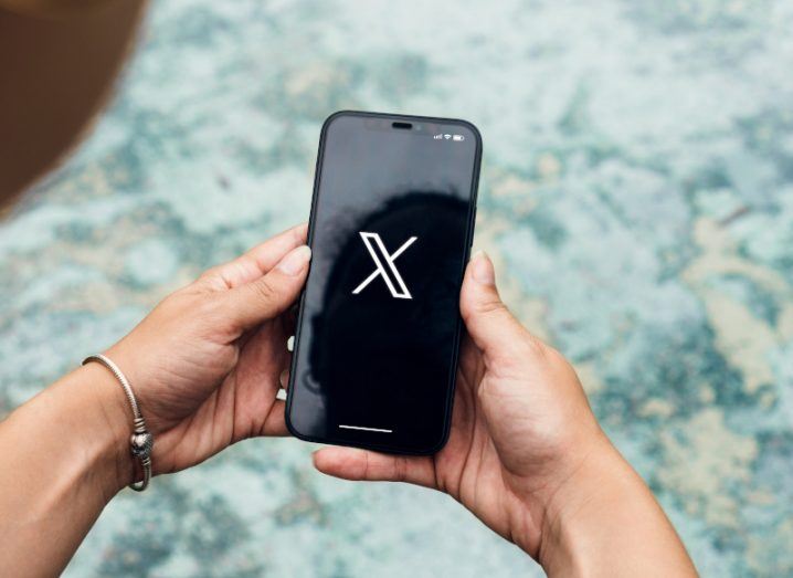 The X logo on a smartphone screen, which is held in a person's hands.