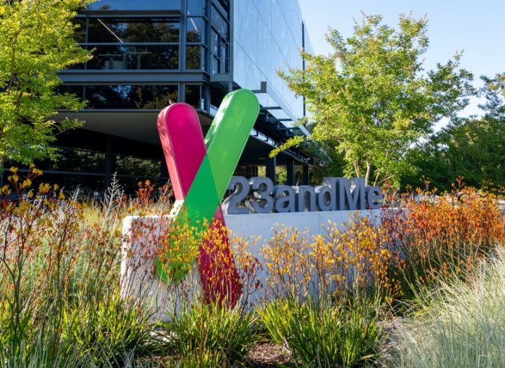 The 23andMe company logo on a sign in a small field, with flowers around it and a building in the background.