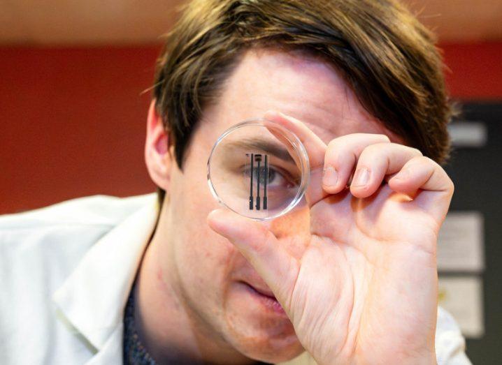 A man in a white lab coat holding a small glass circle in front of his eye. The circle has a small black device in it.