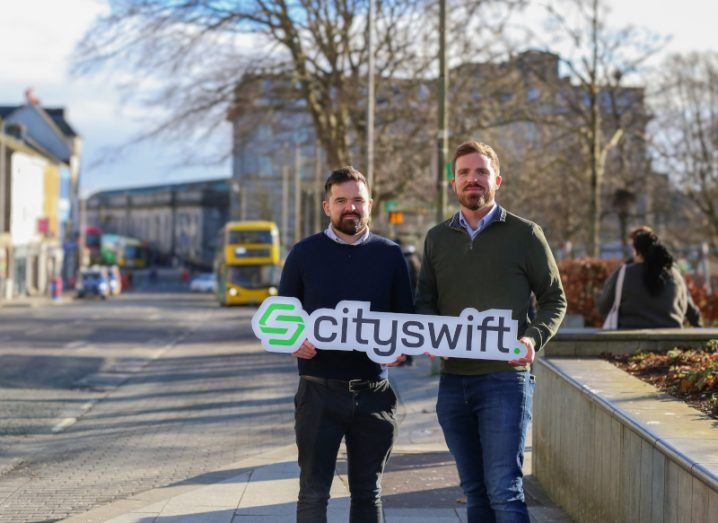 Two men standing on a street in front of a bus holding a CitySwift sign.