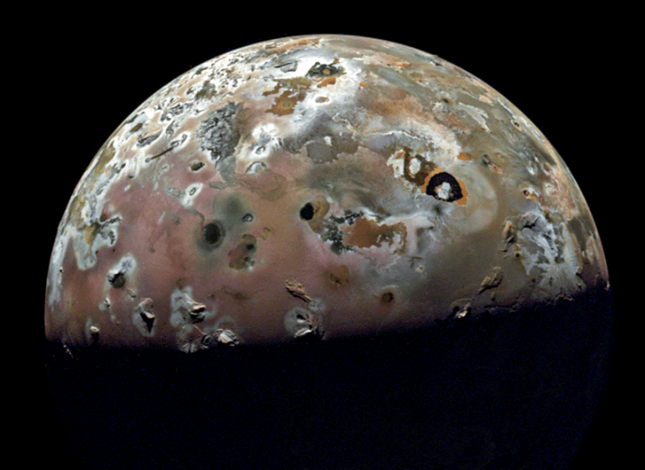 An image of the moon Io, with the lower half of the moon covered in darkness. Different colours are visible on the surface of the moon.
