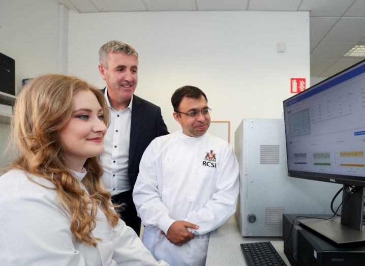 A woman in a white researcher uniform sitting in front of a computer screen in a lab, with two men behind her. One of the men is wearing a similar white researcher outfit. They are part of the RCSI and Serosep.