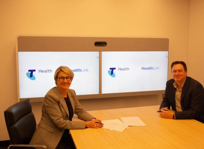 A woman and a man seated at a table with Telstra Health and HealthLink logos on a screen behind them.
