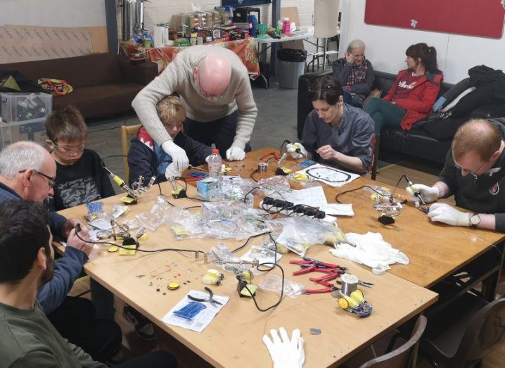 A group of people, including children, sitting around a large table with small hardware devices, learning how to solder at Tog Hackerspace.