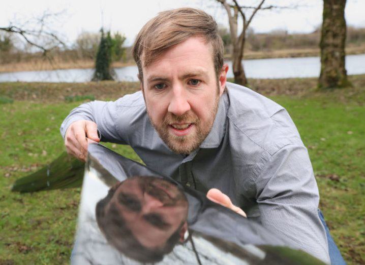 A man standing in front of a reflective turbine foil, with his face visible on the surface of the foil. He is Doctor William Finnegan of the University of Galway.