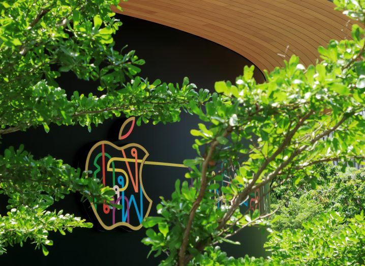 Apple logo in multiple colours on a wall building as soon through some trees.