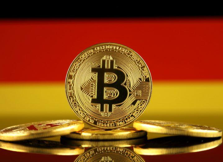 A gold bitcoin sits upright on a stack of other coins in front of a German flag.