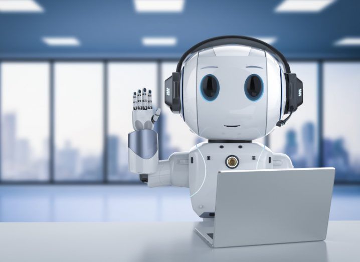 A small, cute robot waves at the camera while sitting at a desk with an open laptop. There is a blurry office and cityscape behind it.
