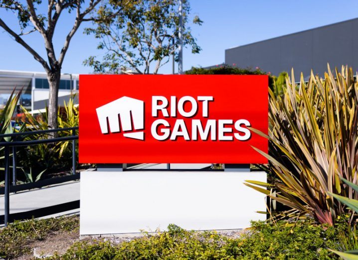 The Riot Games logo on a red sign in front of a building, with a plant next to the sign and a tree in the background.