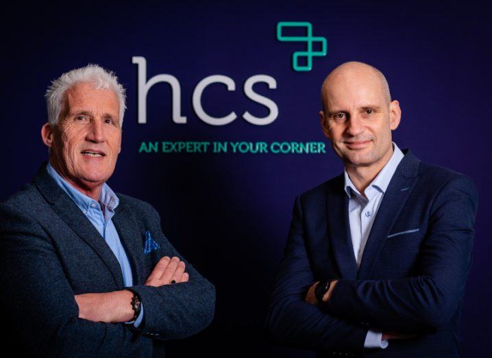 Two men in suits standing next to each other with the HCS company logo on a wall behind them.