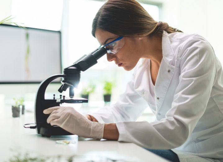 A woman wearing a lab coat, goggles and surgical gloves looks into a microscope in a bright lab setting.