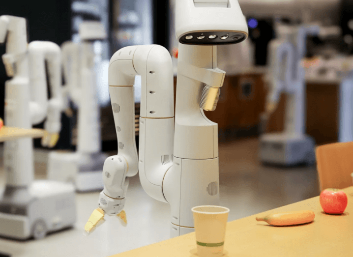 A robotic arm with a camera attachment on the top, above a table that has an apple, a banana and a small cup on it.