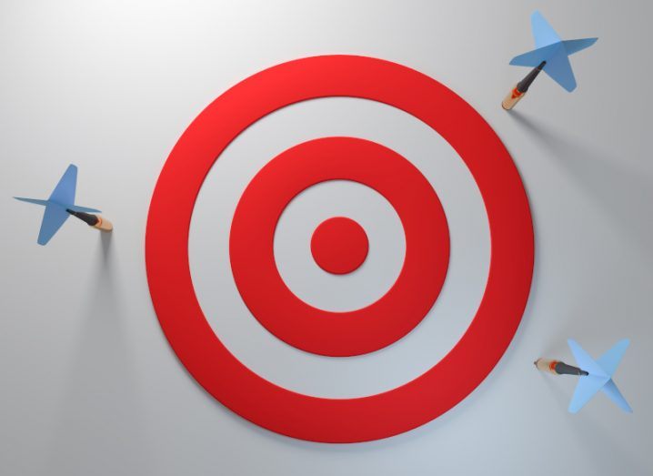 A red target with three blue-headed darts all around the target having missed and stuck to a grey wall behind.