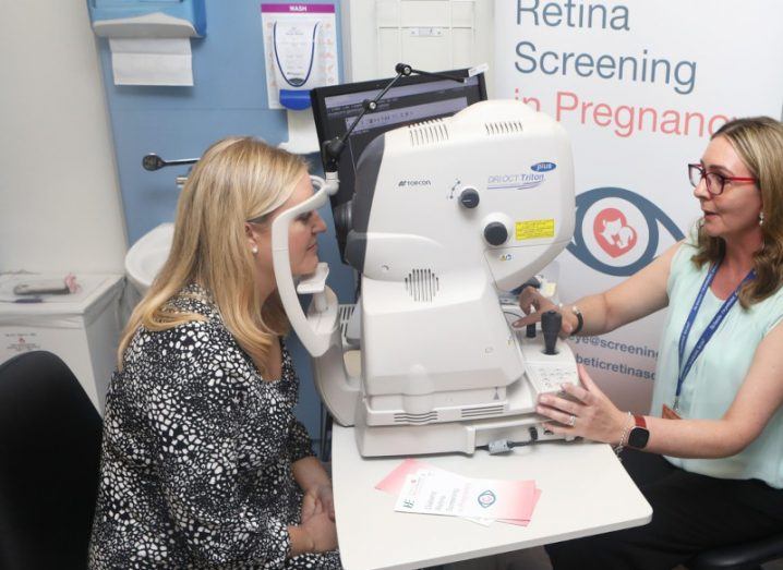 A woman sitting in front of a machine, receiving a diabetic eye screening, while another woman looks at the machine's results.
