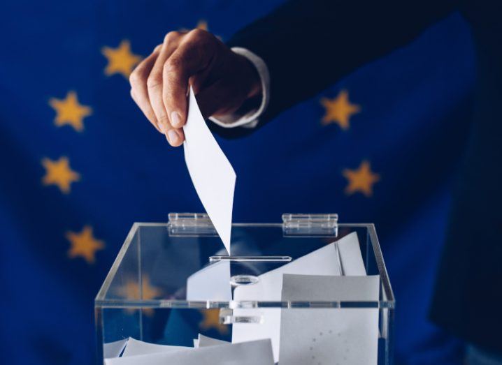 Person putting a vote into a ballot box with an EU flag in the background.