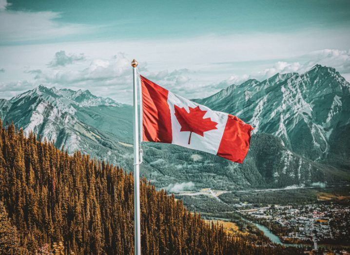 The flag of Canada on a pole with a forest and mountains in the background.