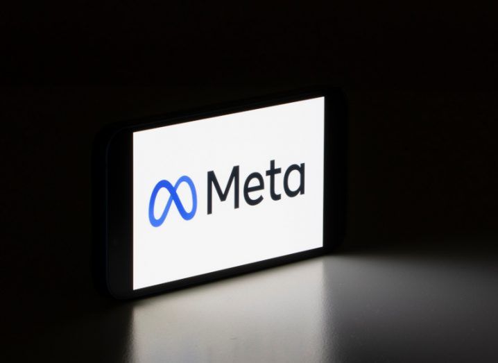 The Meta logo on the front of a smartphone screen, which is on a dark surface with the light from the screen shining out.