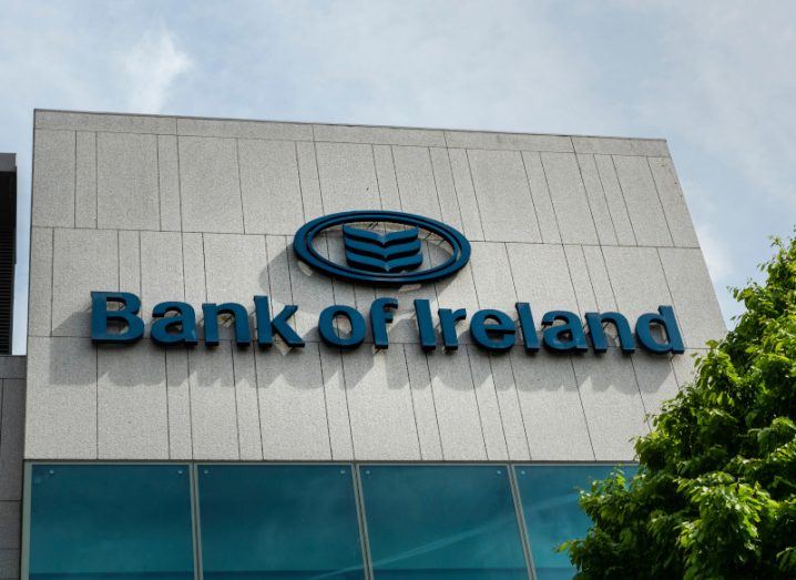 The Bank of Ireland logo on the front of a grey building, with a tree in front of the building and a cloudy sky above.