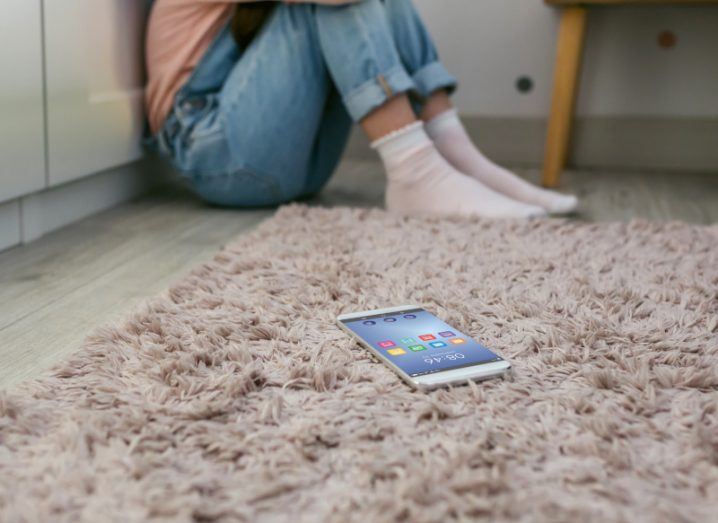 A mobile phone on a rug with a young person sitting by a wall in the background. Used to show the concept of social media impacting the mental health of young people.