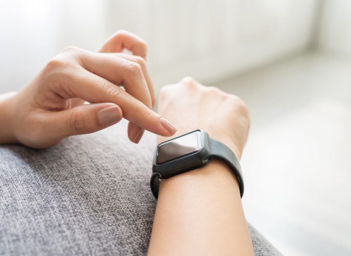 A person touching a smartwatch screen on their wrist. Their wrist is on a couch in a room.