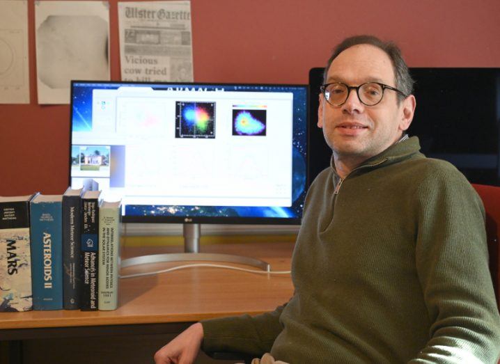 A man wearing glasses and a green jumper sits at a desk with books and a desktop computer on it. He is Doctor Apostolos Christou.