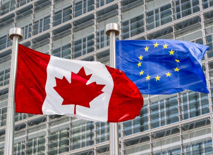 The flags of Canada and the European Union blow in the wind together in front of a high-rise building.