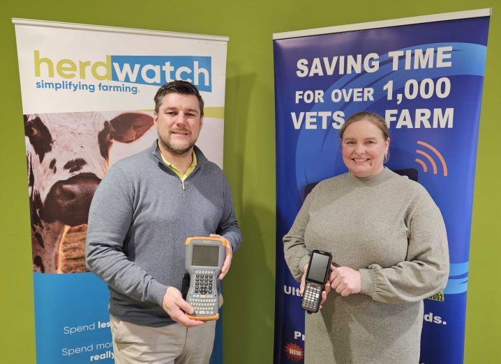 A man and a woman hold farming devices in their hands. Behind them are posters that have the Herdwatch logo on them.