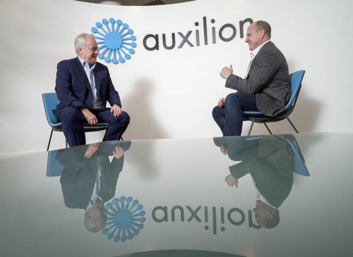 Two men seated in front of each other appearing to talk. Behind them is a big wall with the Auxilion logo on it.