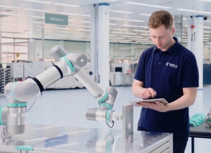 A man wearing a Robco-branded polo shirt working on a robotic arm in a large manufacturing room.