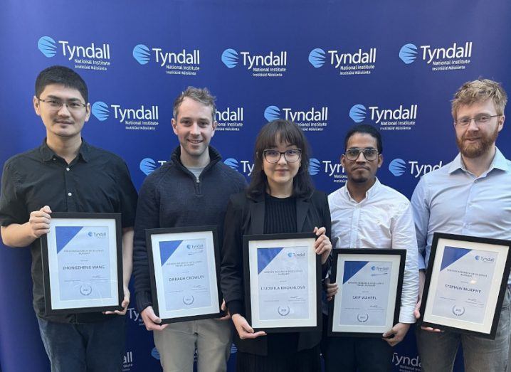 Four men and a woman standing in front of a wall with the Tyndall National Institute logo on it. The five people are holding certificates in front of them, which are Wrixon bursary awards.