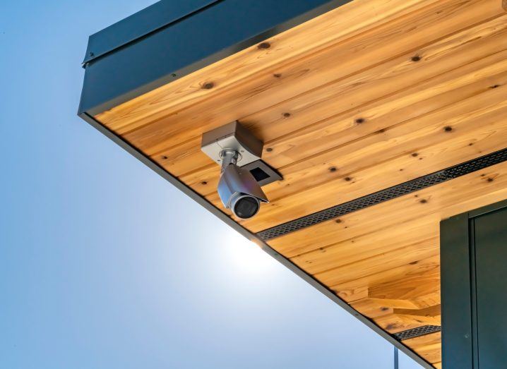 A home security camera on a wooden ceiling with the blue sky and sun in the background.