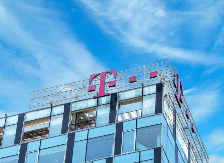 The logo of Deutsche Telekom on a glass building with a blue sky in the backdrop. The logo is a capital T in pin with pink dots around it.