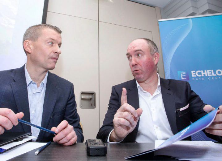 Two men sitting behind a table and wearing suits while speaking to each other. They are part of Echelon Data Centres.