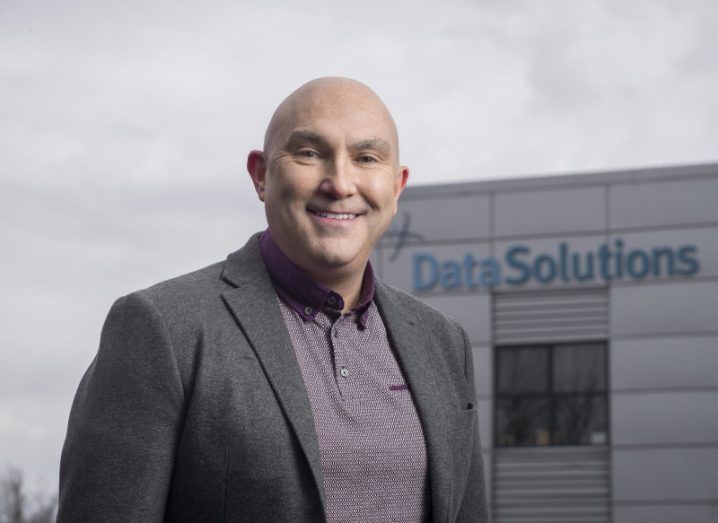 A man wearing a grey suit jacket over a dark purple polo shirt smiles while standing in front of a building. The DataSolutions logo is adorned on the building.