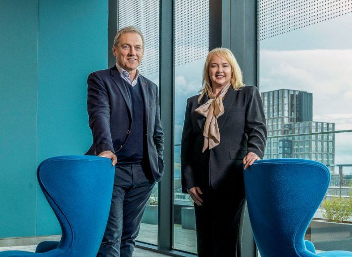 The CEOs of Linked Finance and the Strategic Banking Corporation of Ireland, standing together in front of two blue chairs in an office. A window is behind them, with buildings and a grey sky in the distance.