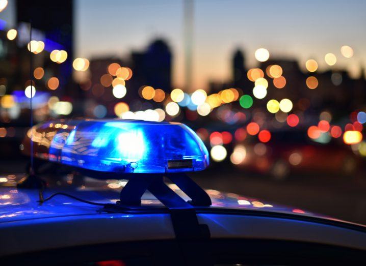 A close-up of the top of a police car with a flashing blue light on with a city in the background at dusk.