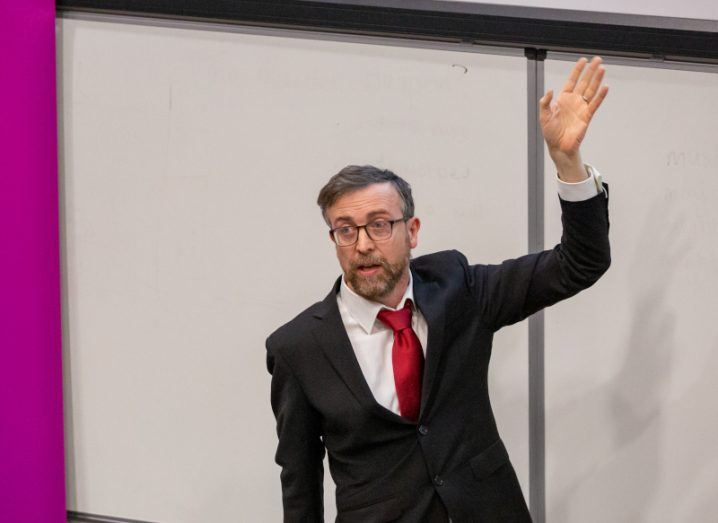A photo of Tomas Ward standing in front of a whiteboard delivering a lecture. One of his arms is raised in a gesture as he speaks. He is wearing a dark suit, white shirt and pink tie.