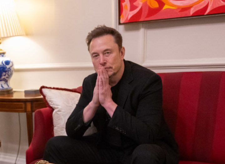 Elon Musk sitting on a red chair with his hands in front of his face.