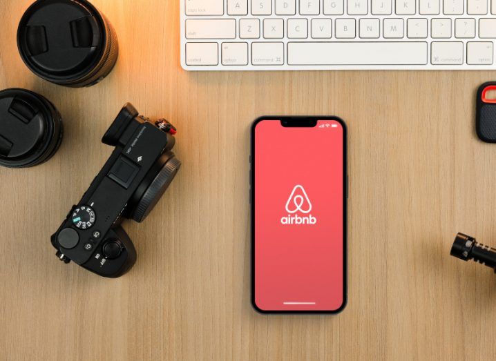 The Airbnb logo on the screen on a smartphone, which is laying on a wooden desk next to a camera and a keyboard.