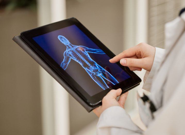 A medical researcher wearing a white lab coat holding a tablet with a 3D scan rendering of a human body visible on the screen.