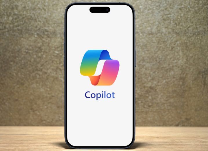 The Microsoft Copilot logo on the screen of a smartphone that is on a wooden table.