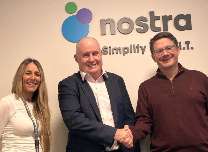 Two men and a woman standing together in front of a wall with the Nostra logo on it.